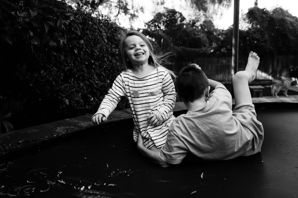 black and white image of a little girl laughing as she falls down on a trampoline; her brother