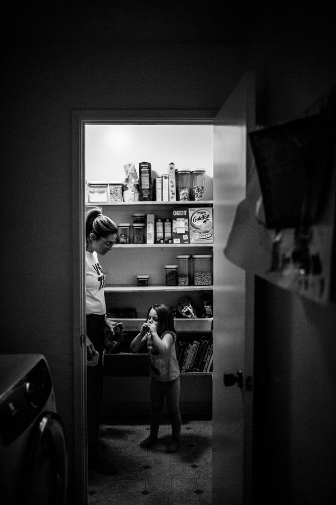 black and white image of a mom with her hair in a ponytail helping her young daughter get a snack out of the pantry