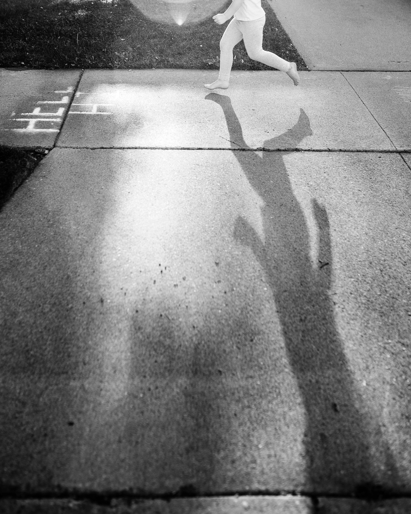 black and white photograph of a little girl skipping along a sidewalk with her shadow