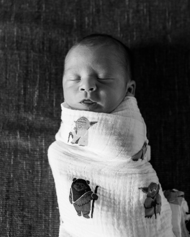 black and white image of a newborn baby boy with his mouth slightly open, wrapped in a Yoda and Ewok swaddle