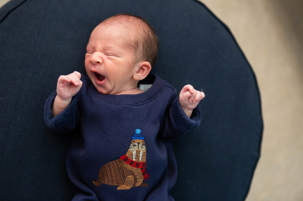 yawning baby boy wearing a navy onesie with an embroidered walrus wearing a hat and scarf